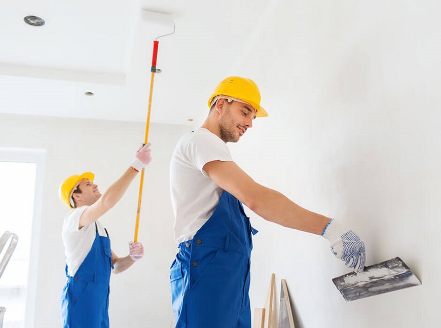 Two workers in blue overalls and yellow hard hats apply material to a bright white wall, with one using a long-handled roller and the other a trowel; bright indoor lighting.