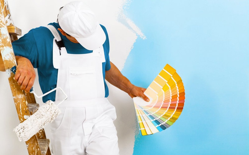 A worker in white overalls holds a fan of color samples and a paint roller beside a partially blue-painted wall; bright studio lighting.